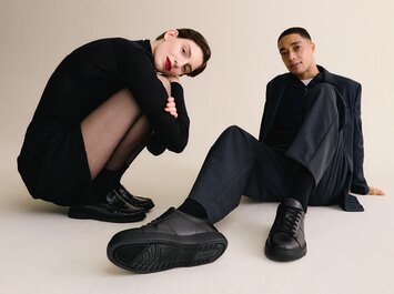 A woman and a man sit relaxed on the floor, both wearing dark, elegant outfits and black Gabor shoes, whose soles are clearly visible in the foreground. | © Gabor Shoes GmbH, Germany